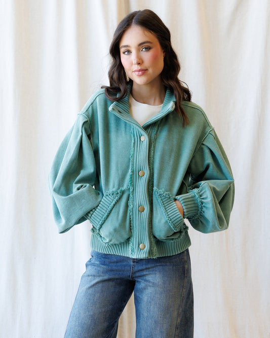 A woman wearing a sage green ribbed knit button-up cardigan with voluminous balloon sleeves and front pockets, paired with blue jeans, standing against a draped cream fabric backdrop.