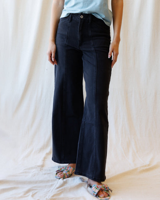 A person wearing dark navy wide-leg denim pants with a light blue top and patterned flat sandals, standing against a draped cream fabric backdrop.