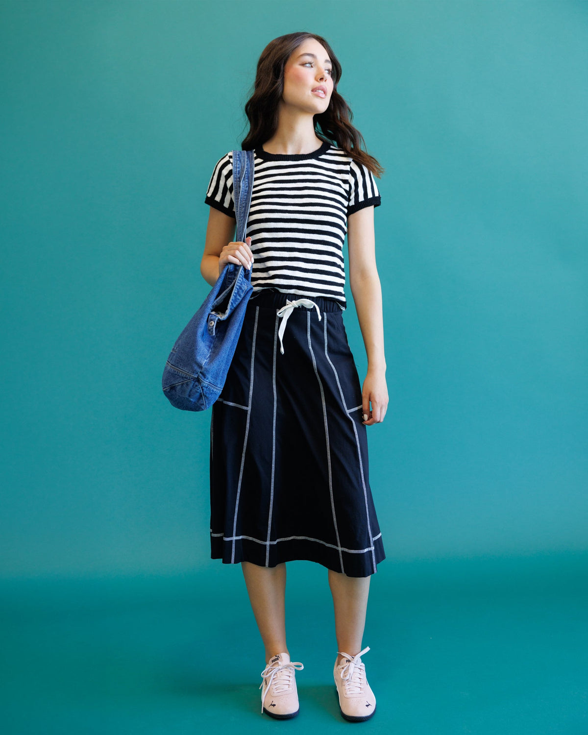 A woman modeling a black and white striped short-sleeve top with a navy blue midi skirt featuring white piping and a white drawstring, holding a blue tote bag against a solid teal background.