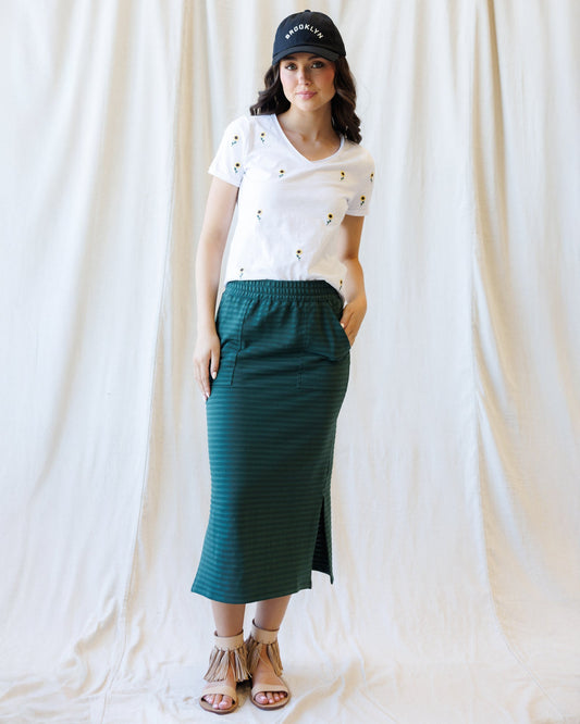 A woman stands against a draped white fabric backdrop wearing a white short-sleeve V-neck T-shirt with small embroidered details, a dark green striped midi skirt with side slits, a black baseball cap, and tan strappy sandals.