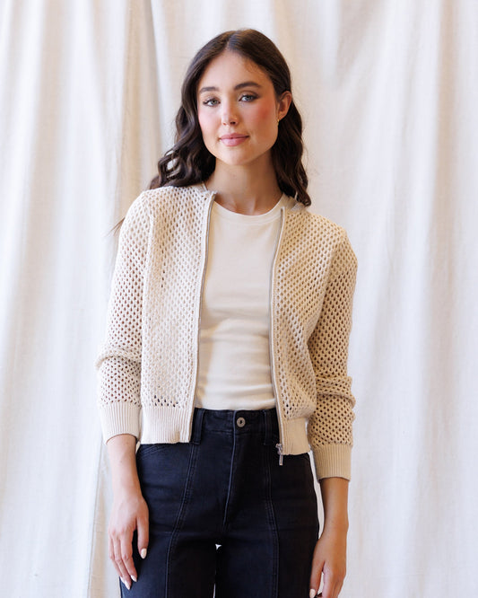 A woman modeling a cream-colored open-knit zip-up bomber jacket over a matching cream t-shirt and dark blue jeans, standing against a draped white fabric backdrop.