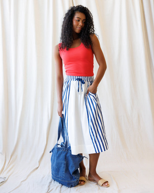Woman wearing a red top and blue and white striped skirt, holding a denim bag against a white curtain background.