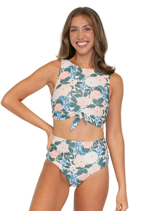 A woman modeling a two-piece floral bikini set against a plain white background. The set features a high-neck crop top with a front tie detail and matching high-waisted bottoms, both in a green, pink, and blue floral pattern.
