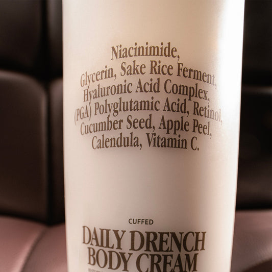 Close-up of a bottle of 'Daily Drench Body Cream' with a list of ingredients on a blurred background.