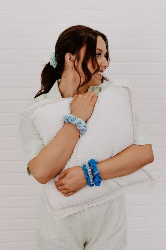 A woman wearing a light green gingham pajama set and matching scrunchies holds a white textured pillow against her chest, posing in front of a white brick wall.