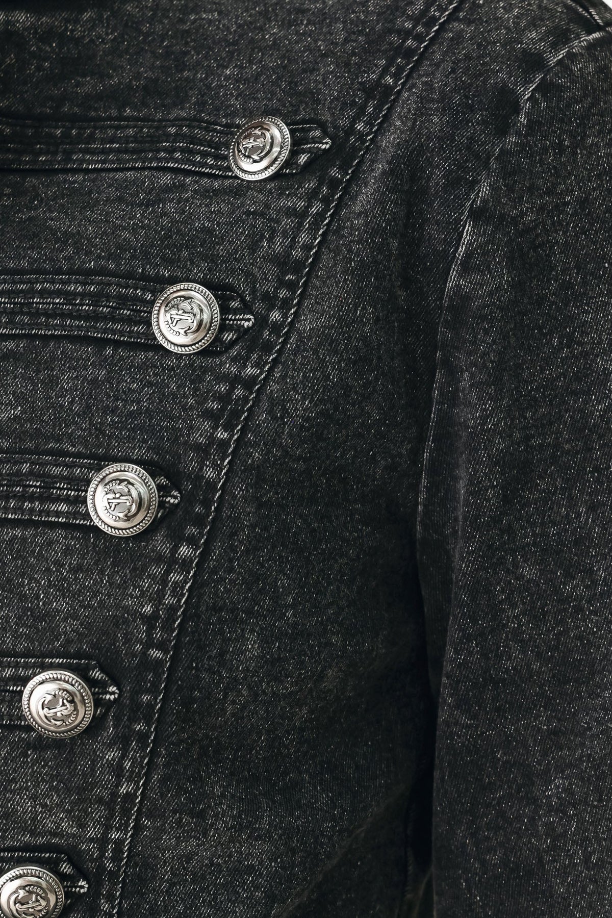 Close-up of a black washed denim jacket featuring a row of decorative silver-tone metal buttons with a crest medallion.