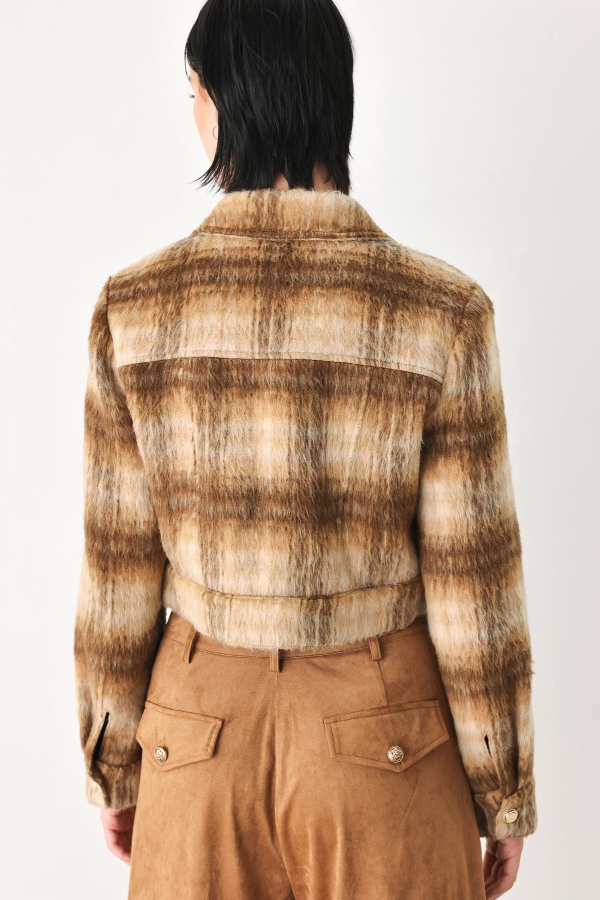A model shown from the back wearing a cropped, long-sleeve plaid jacket in brown and beige tones, paired with brown trousers, against a plain white studio background.