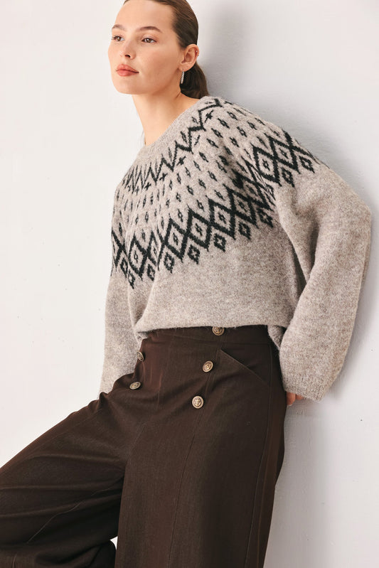 A woman wearing a light gray knit sweater with a black geometric pattern across the chest and shoulders, paired with dark brown high-waisted trousers, posing against a plain off-white wall.