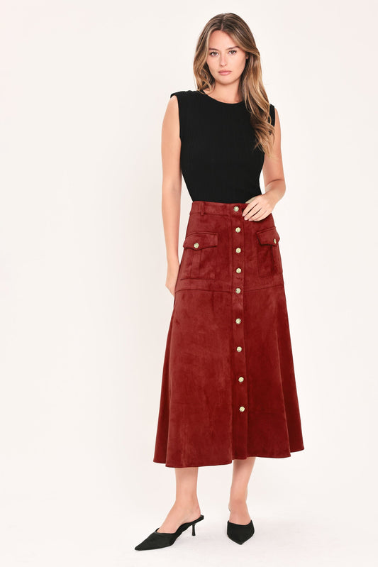 A woman modeling a high-waisted, mid-length rust red corduroy skirt with gold-tone button details down the front, paired with a black sleeveless top and black pointed-toe mule heels, against a plain off-white studio background.