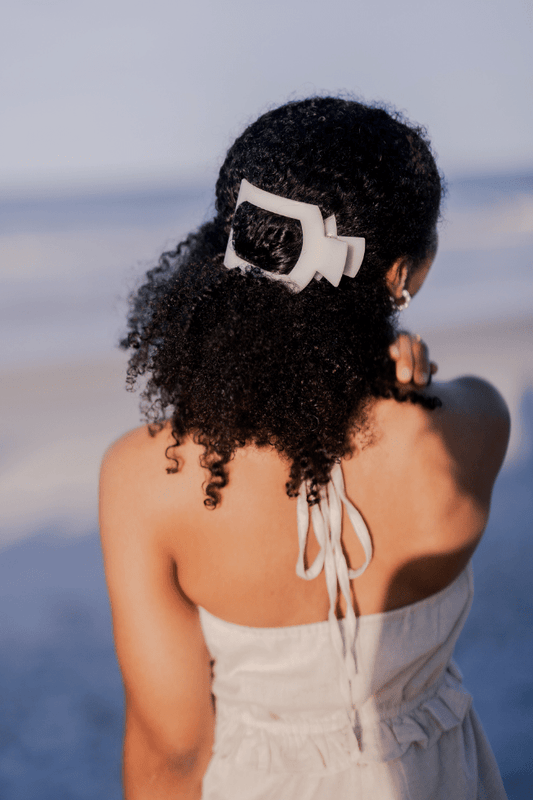 Woman with a white hair clip in her curly hair, standing on a beach.