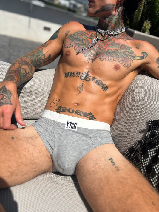 A tattooed man reclining outdoors on a light-colored lounge chair, wearing heather gray boxer briefs with a white waistband and a visible YYCG logo.