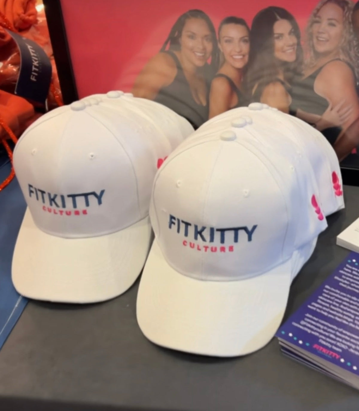 Two white caps with 'FITKITTY Culture' text on a table with promotional material in the background.