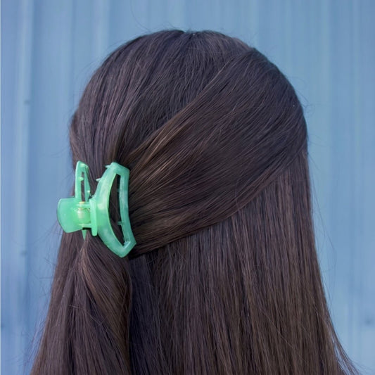 A mint green plastic claw clip holding back a section of dark brown hair, shown from the back against a light blue background.