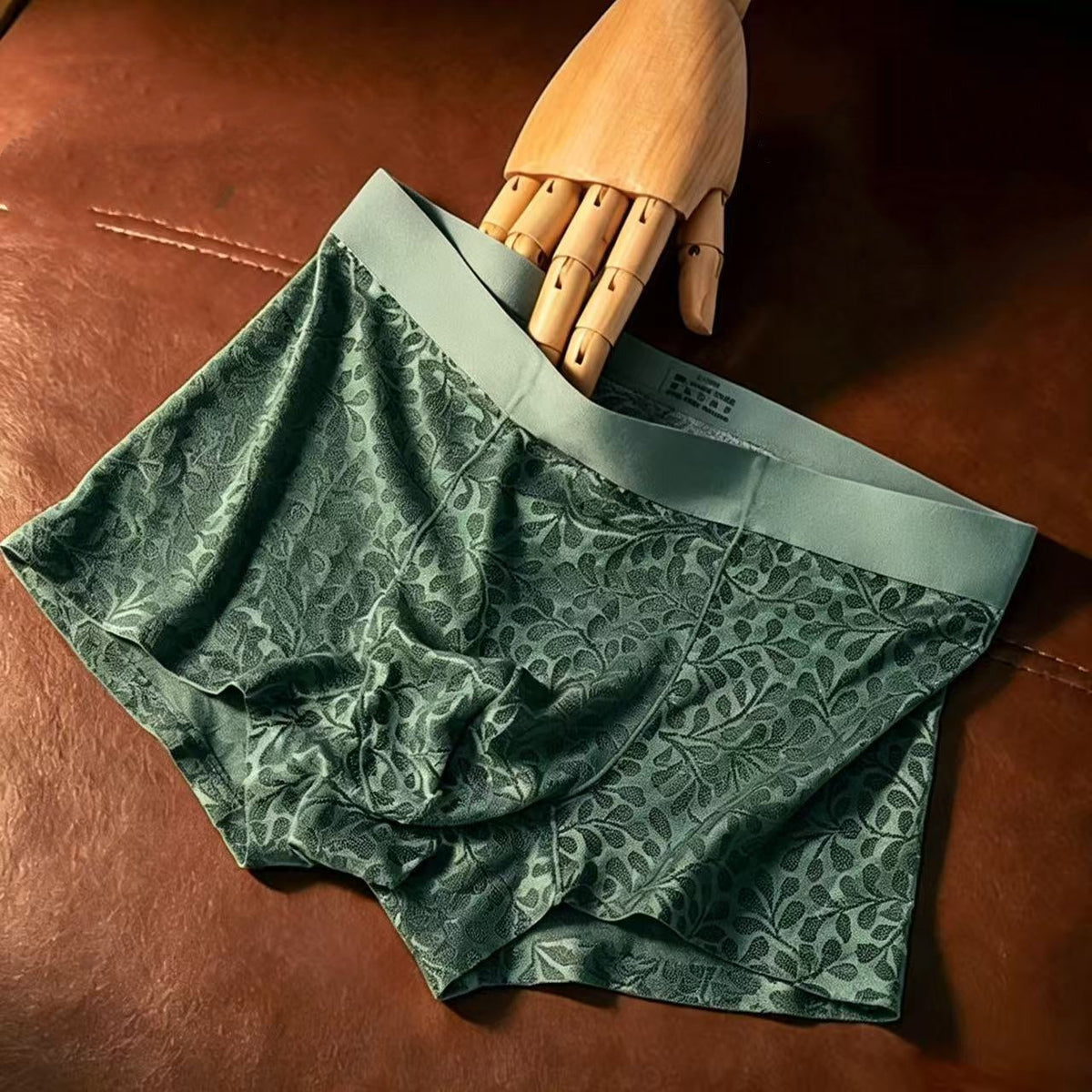 A pair of men's boxer briefs in a muted green floral jacquard pattern with a solid sage green waistband, displayed on a brown leather surface with a wooden mannequin hand holding the waistband.