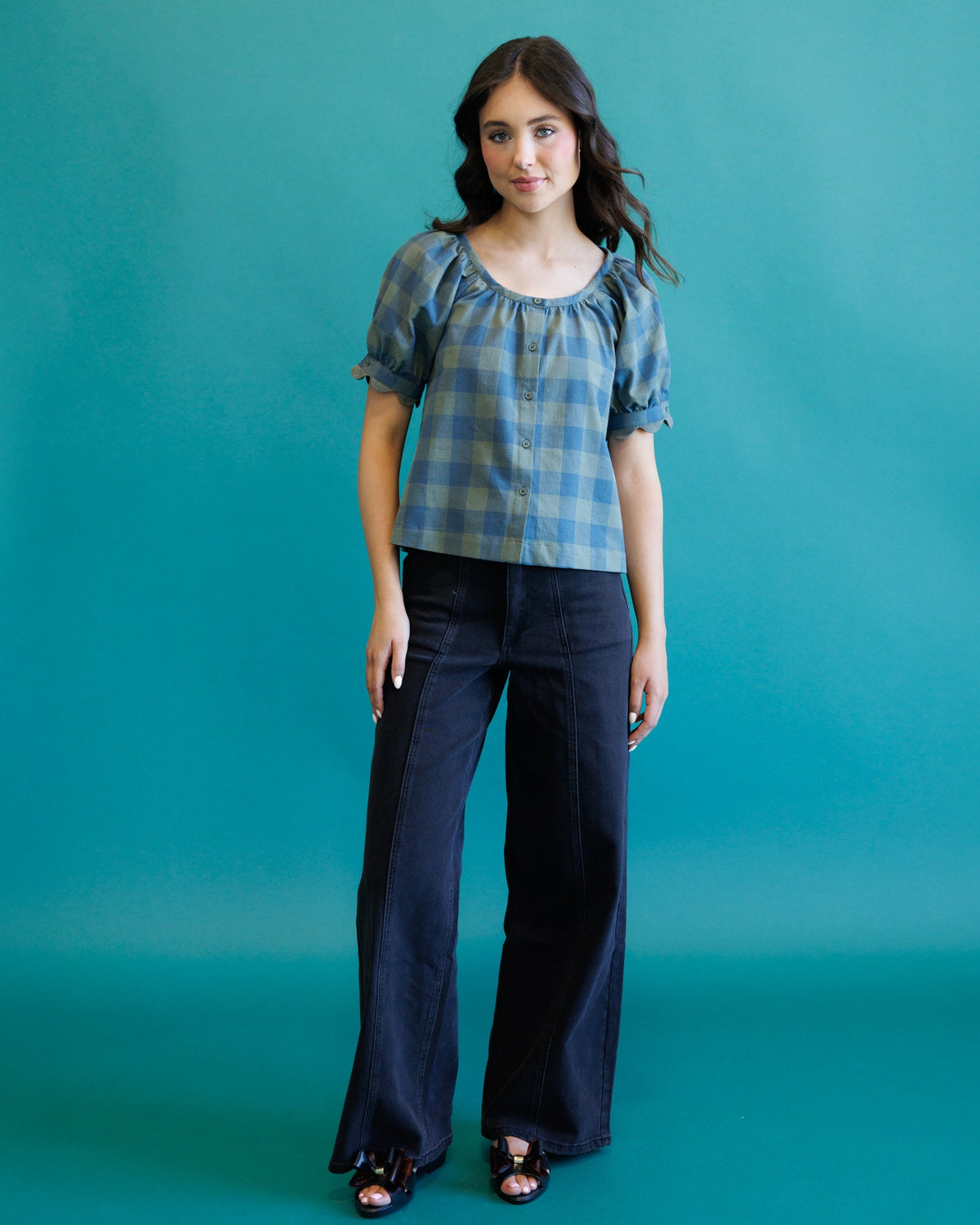 A woman modeling a blue and gray plaid puff-sleeve top paired with dark wide-leg pants, standing against a solid teal background.