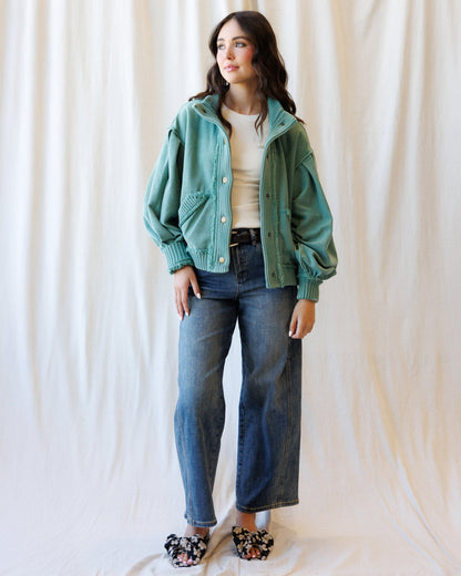 A woman stands against a draped cream fabric backdrop wearing a sage green corduroy jacket over a cream top, blue wide-leg jeans, and black floral slip-on shoes.
