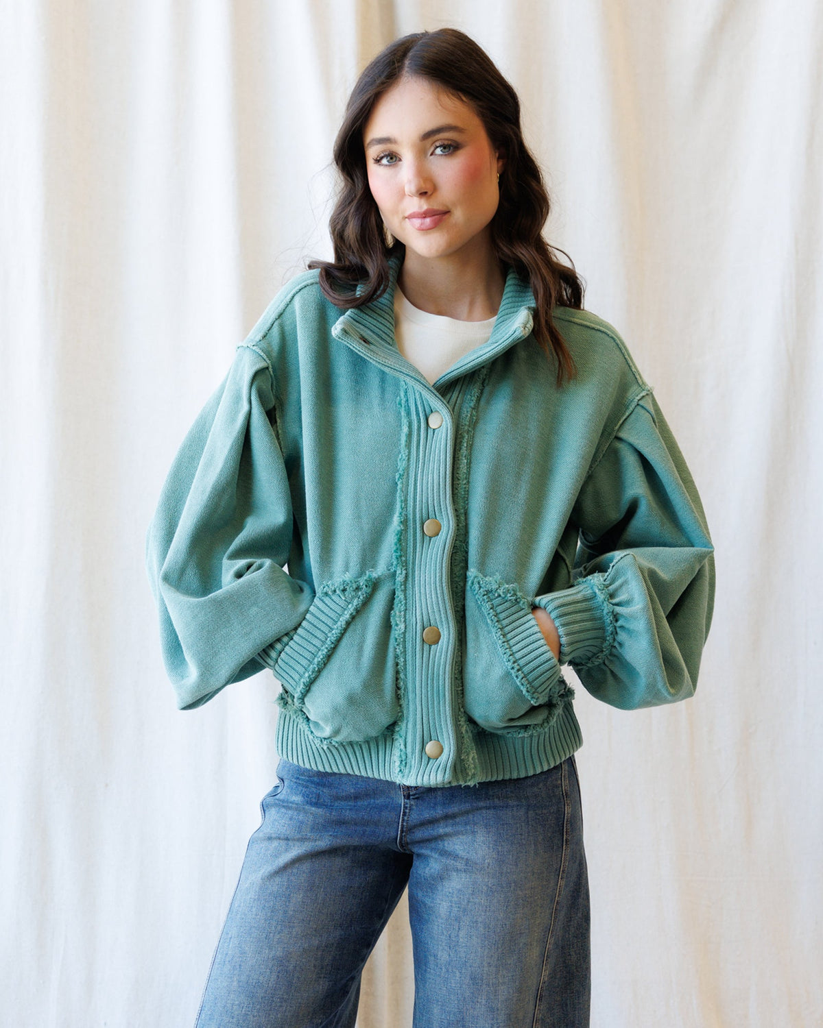 A woman wearing a sage green ribbed knit button-up cardigan with voluminous balloon sleeves and front pockets, paired with blue jeans, standing against a draped cream fabric backdrop.