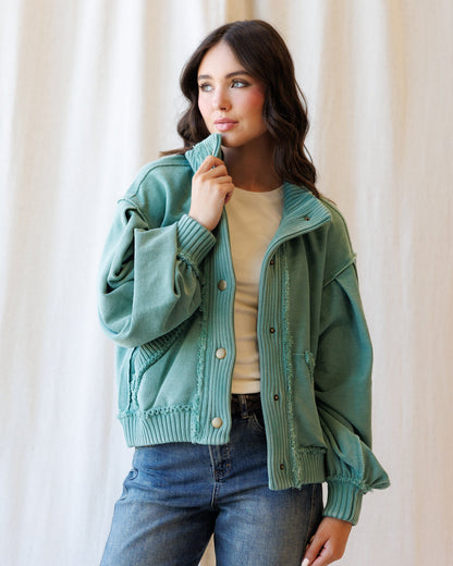 A woman wearing a sage green ribbed knit cardigan jacket over a beige top and blue jeans, standing against a soft beige curtain background.