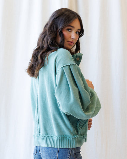 A woman wearing a sage green knit sweater with a high mock neck and ribbed hem, paired with blue jeans, standing against a soft white curtain background.