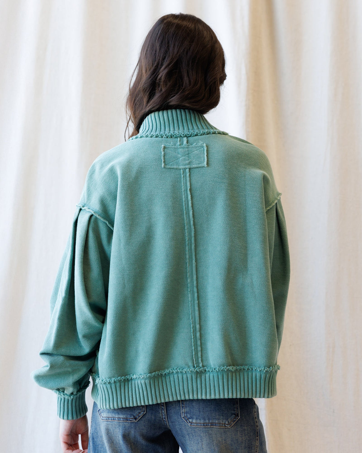 A person viewed from the back wearing a mint green knit sweater with a mock neck, dropped shoulders, and ribbed cuffs and hem, paired with blue jeans against a cream curtain backdrop.