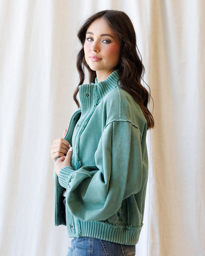 A woman wearing a sage green ribbed knit cardigan sweater with a mock neck and button front, paired with blue jeans, standing against a soft beige curtain backdrop.
