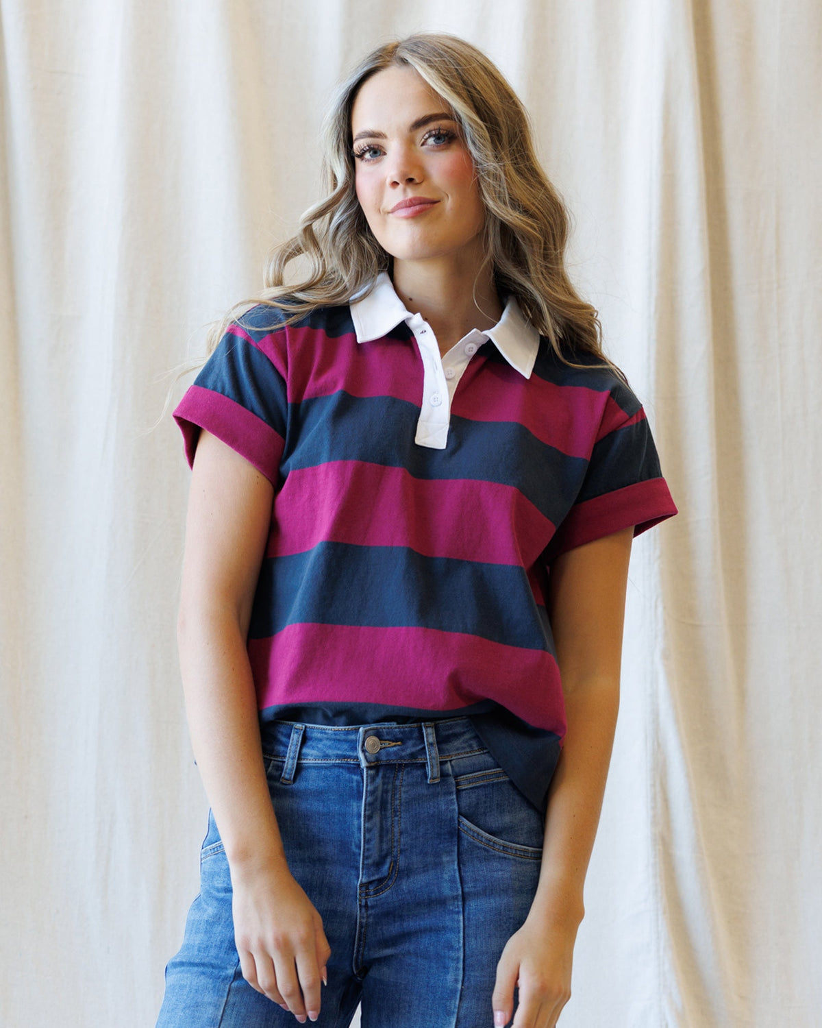 A woman wearing a short-sleeve polo shirt with horizontal stripes in navy blue, magenta, and dark gray, paired with blue jeans, standing against a draped cream fabric backdrop.