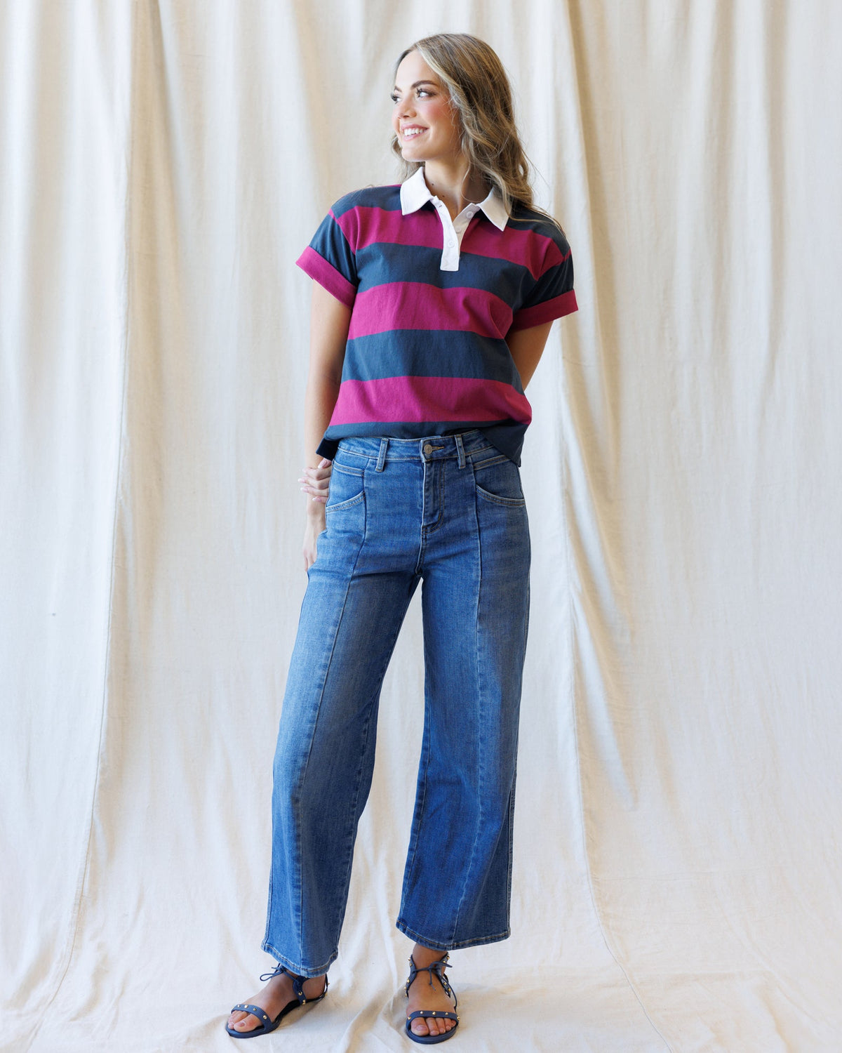 A woman modeling a short-sleeve polo shirt with horizontal navy, magenta, and dark gray stripes, paired with high-waisted blue wide-leg jeans and brown sandals, standing against a draped cream fabric backdrop.