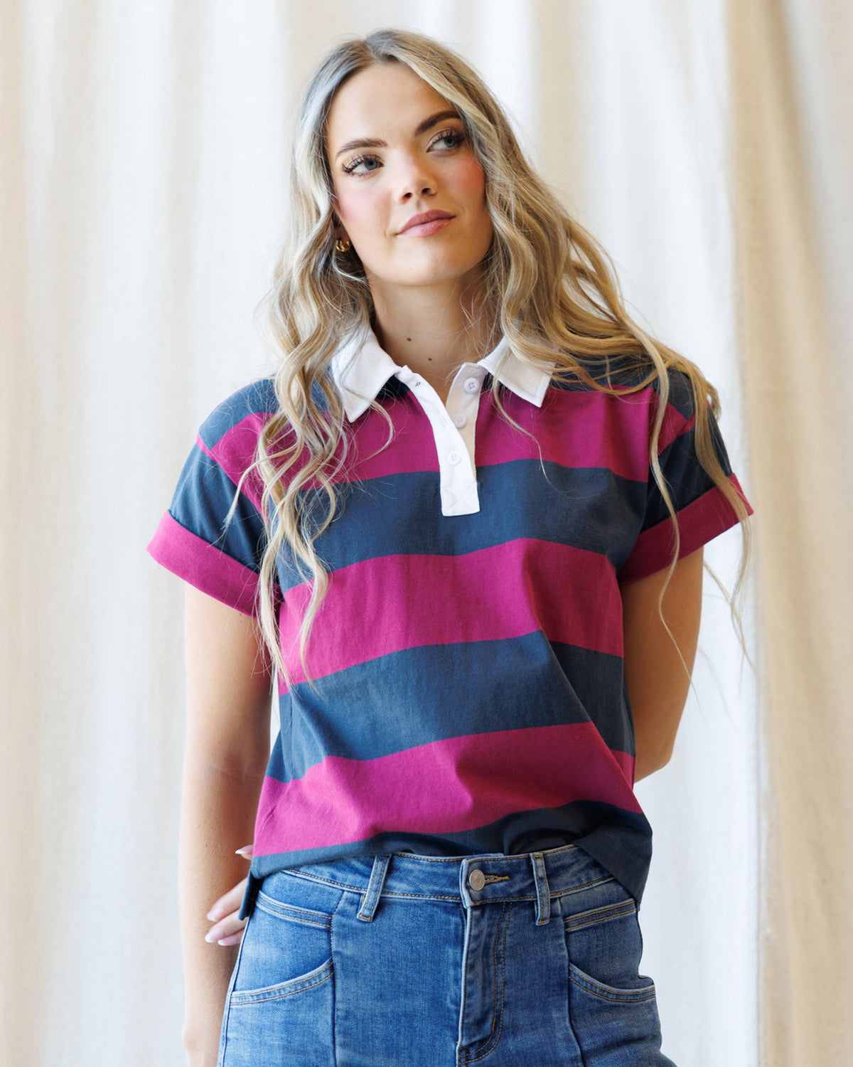 A woman wearing a short-sleeve polo shirt with horizontal stripes in navy blue, magenta pink, and white, paired with blue jeans, standing against a soft beige curtain background.