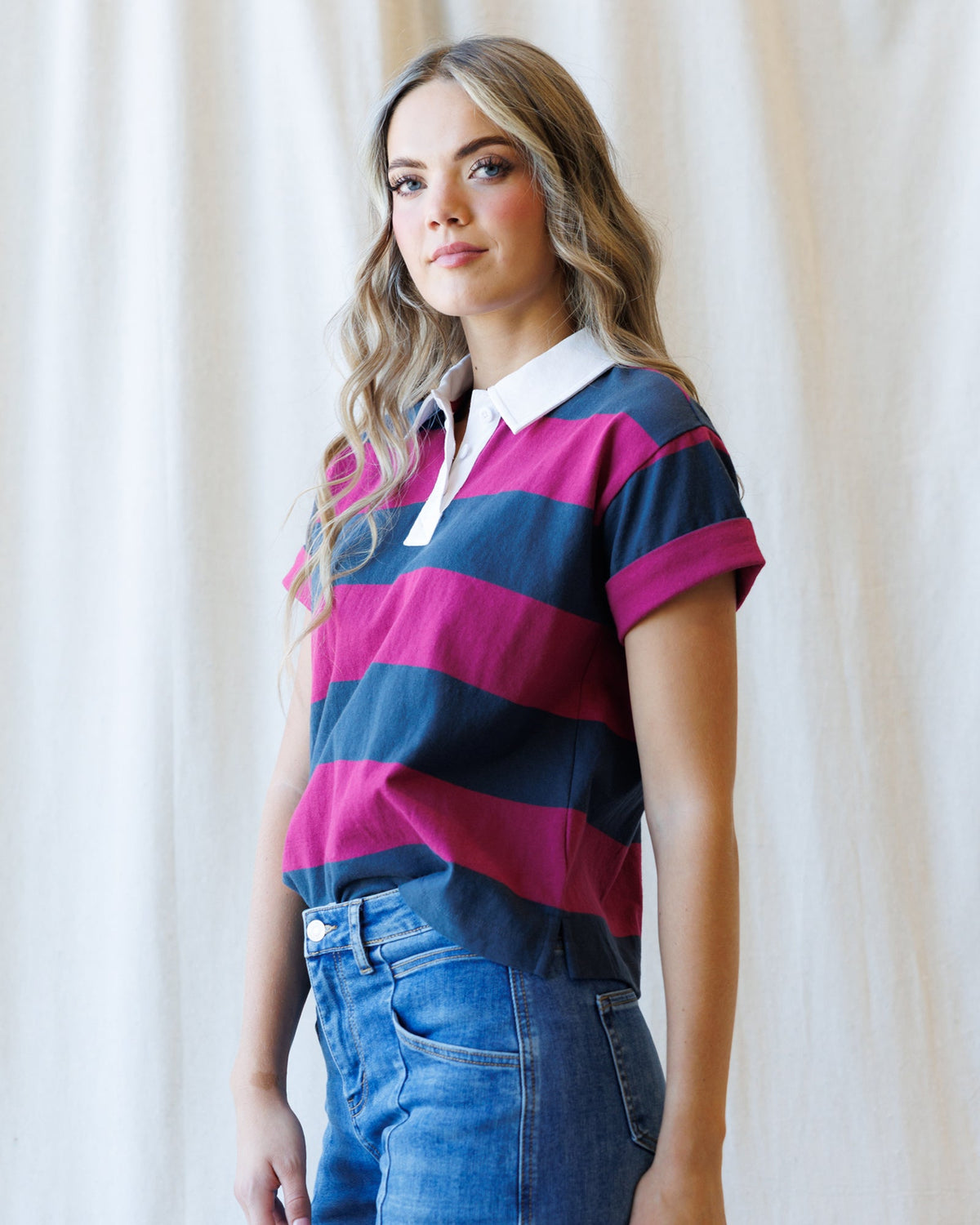 A woman wearing a short-sleeve polo shirt with bold magenta and navy blue horizontal stripes, paired with blue jeans, standing against a soft beige curtain backdrop.