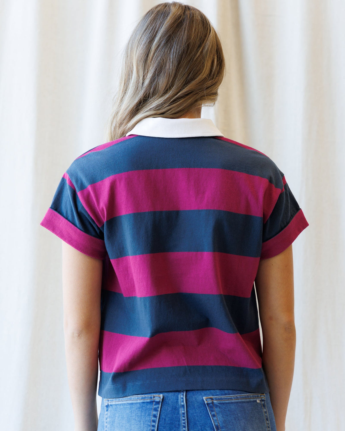 A woman viewed from behind wearing a short-sleeve polo shirt with bold horizontal navy blue and magenta pink stripes, paired with blue jeans against a soft cream curtain background.
