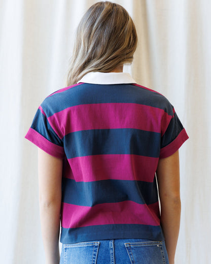 A woman viewed from behind wearing a short-sleeve polo shirt with bold horizontal navy blue and magenta pink stripes, paired with blue jeans against a soft cream curtain background.