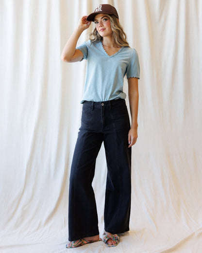 A woman modeling a light blue short-sleeve V-neck top with scalloped edges, paired with dark wide-leg jeans, a brown baseball cap, and flat sandals, standing against a draped cream fabric backdrop.
