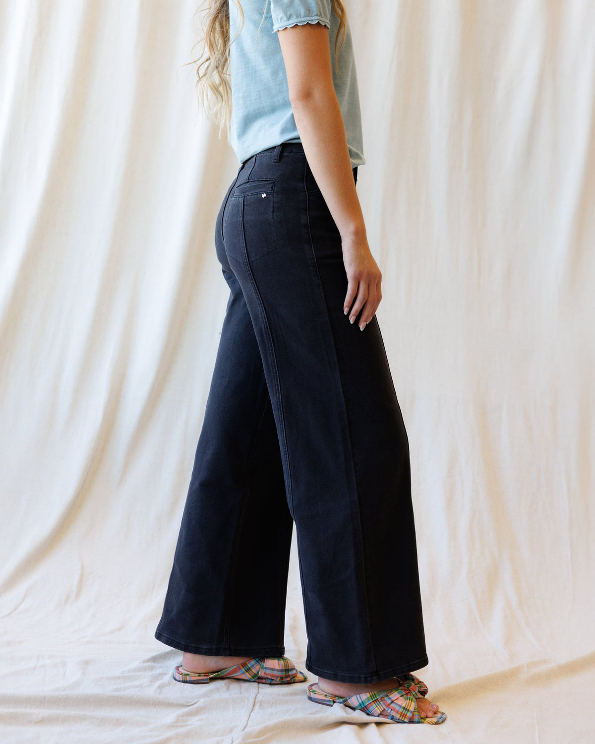 A person shown from the waist down wearing dark wide-leg denim pants, a light blue t-shirt, and colorful plaid slide sandals, standing against a draped cream fabric backdrop.
