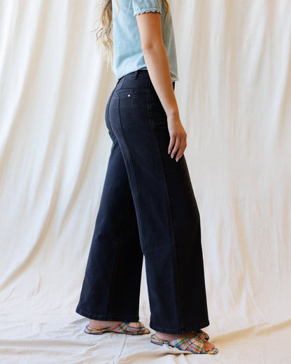 A person shown from the waist down wearing dark wide-leg denim pants, a light blue t-shirt, and colorful plaid slide sandals, standing against a draped cream fabric backdrop.