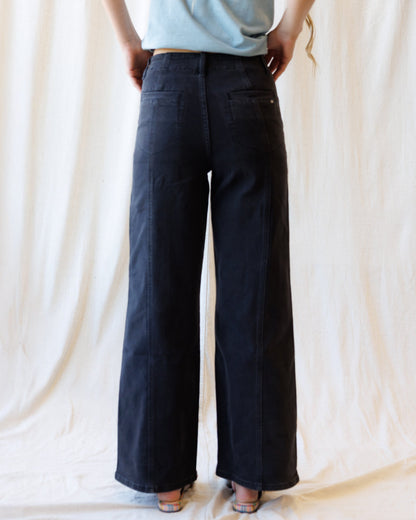 A person shown from the back wearing dark wide-leg pants and a light blue top, standing against a draped cream fabric backdrop.