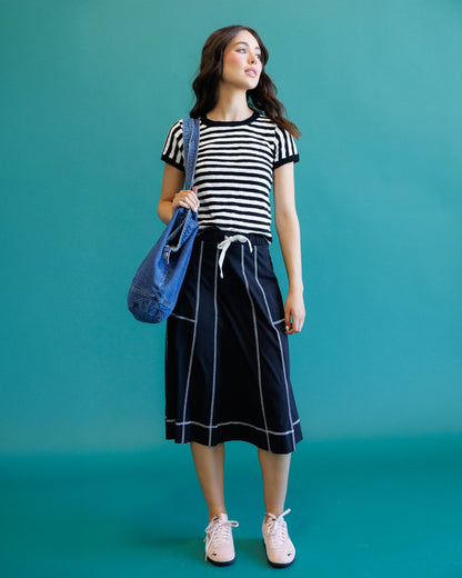 A woman modeling a black and white striped short-sleeve top with a navy blue midi skirt featuring white piping and a white drawstring, holding a blue tote bag against a solid teal background.