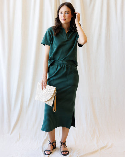 A woman modeling a dark green two-piece set consisting of a short-sleeve collared top and a matching midi skirt with a side slit, styled with a white woven clutch and black strappy sandals against a draped white fabric backdrop.