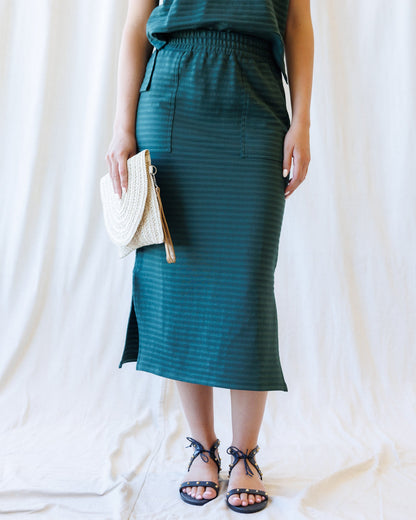 A model wearing a dark teal green striped midi skirt with a side slit, holding a woven cream clutch, and wearing black strappy sandals against a draped white fabric backdrop.