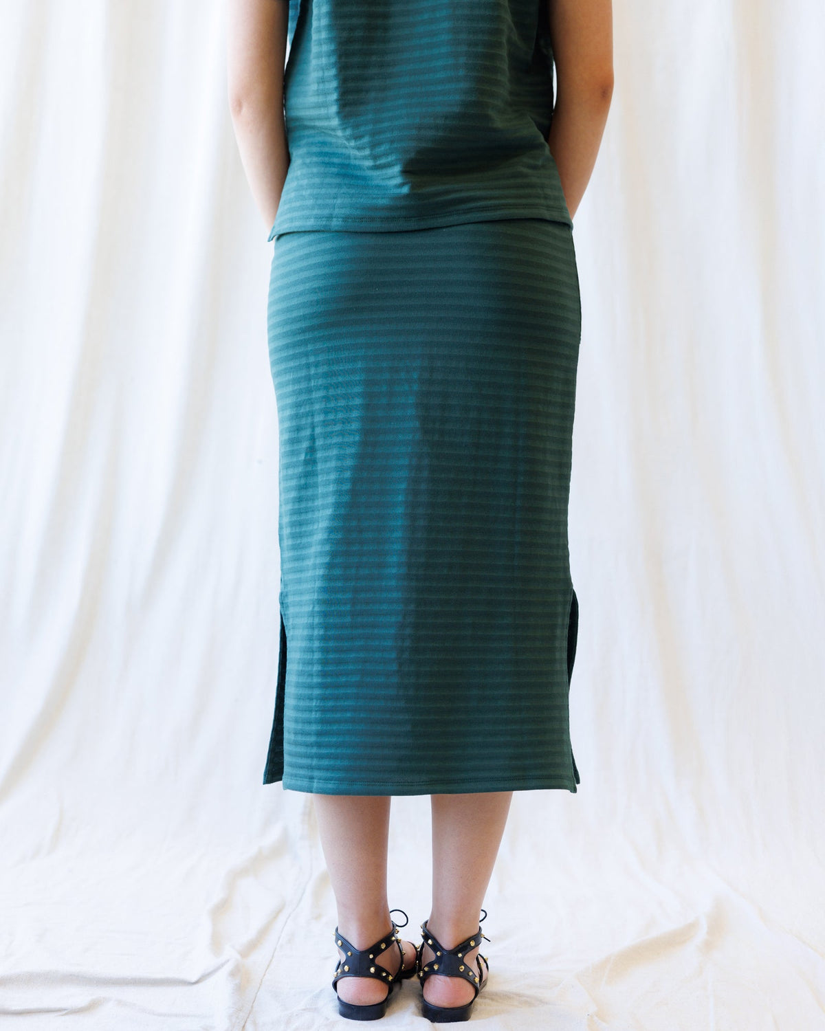 A person shown from the back wearing a dark teal green ribbed midi skirt with side slits and a matching top, paired with black lace-up sandals, set against a draped white fabric backdrop.