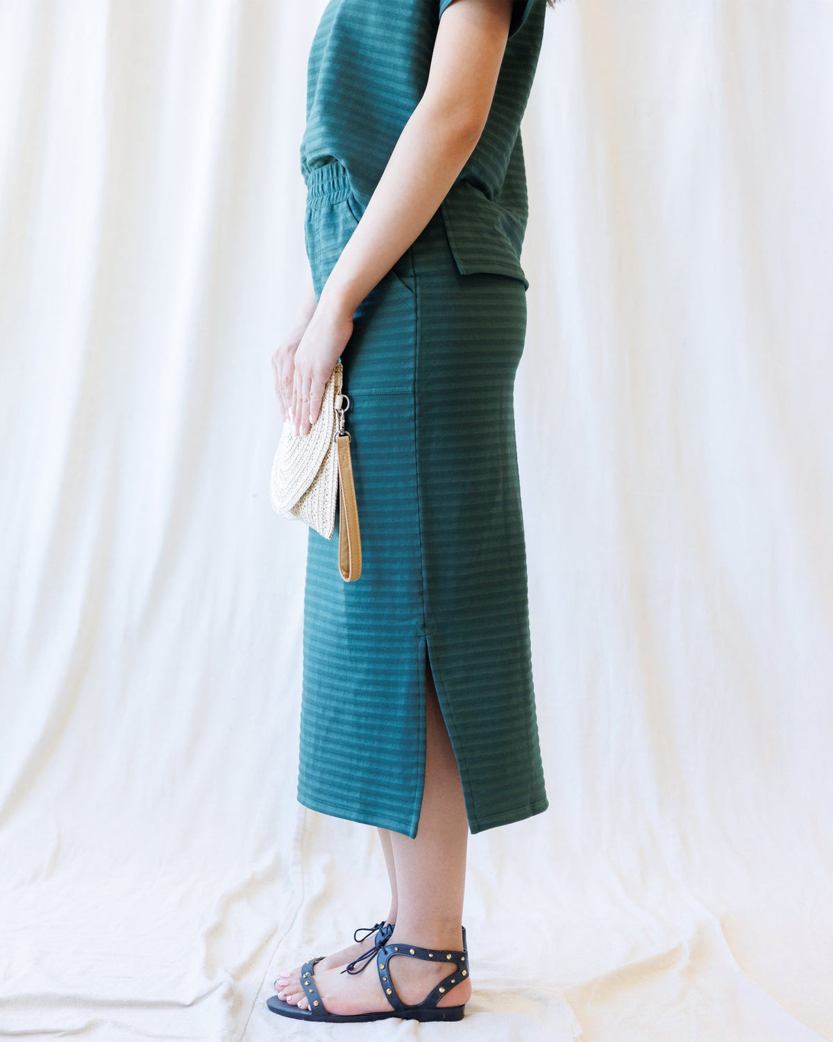 A person wearing a dark teal green striped midi dress with a side slit, holding a woven cream clutch, standing against a draped white fabric backdrop.