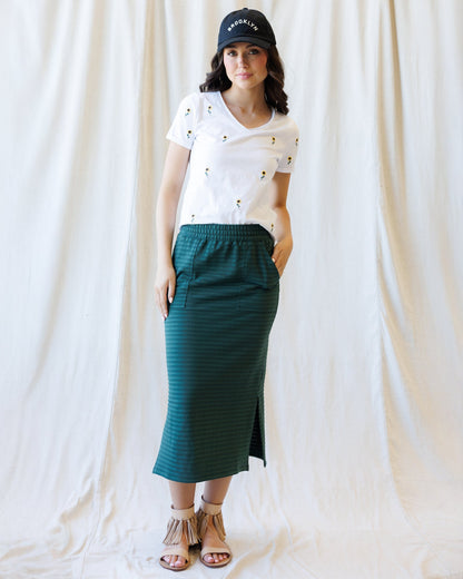 A woman stands against a draped white fabric backdrop wearing a white short-sleeve V-neck T-shirt with small embroidered details, a dark green striped midi skirt with side slits, a black baseball cap, and tan strappy sandals.