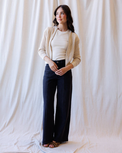 A woman stands against a draped white fabric backdrop wearing a cream knit cardigan over a cream top, dark navy wide-leg pants, and leopard-print flat sandals.