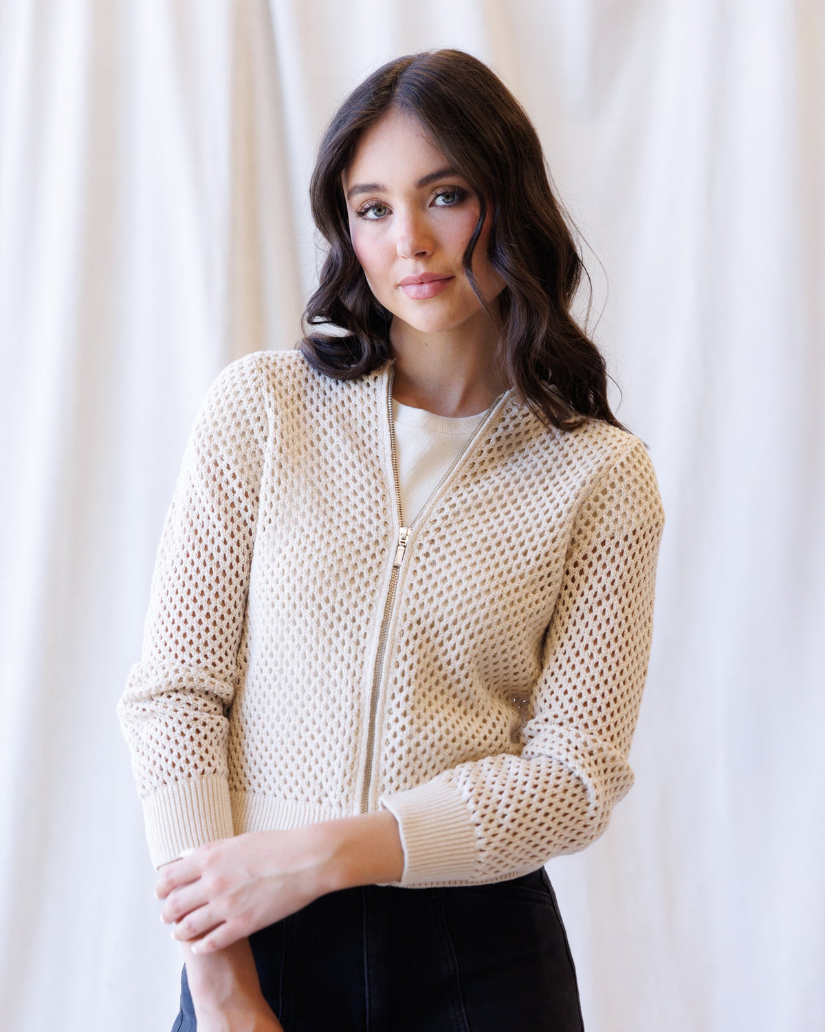 A woman wearing a cream-colored open-knit zip-up cardigan over a white top and black pants, standing against a soft white curtain background.