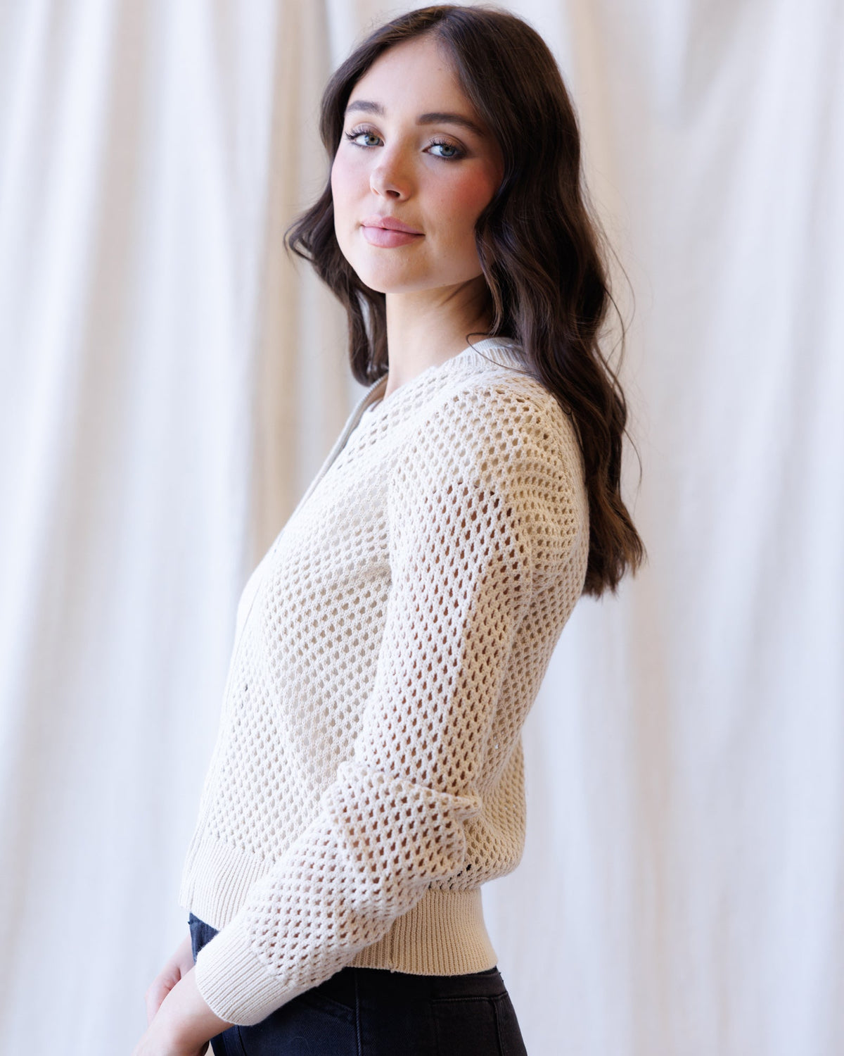 A woman with long dark brown hair is shown from the side, wearing a cream-colored open-knit long-sleeve sweater and dark pants, standing against a soft white curtain background.