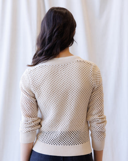 A woman viewed from behind wearing a cream-colored open-knit sweater with ribbed cuffs and hem, set against a soft white curtain background.