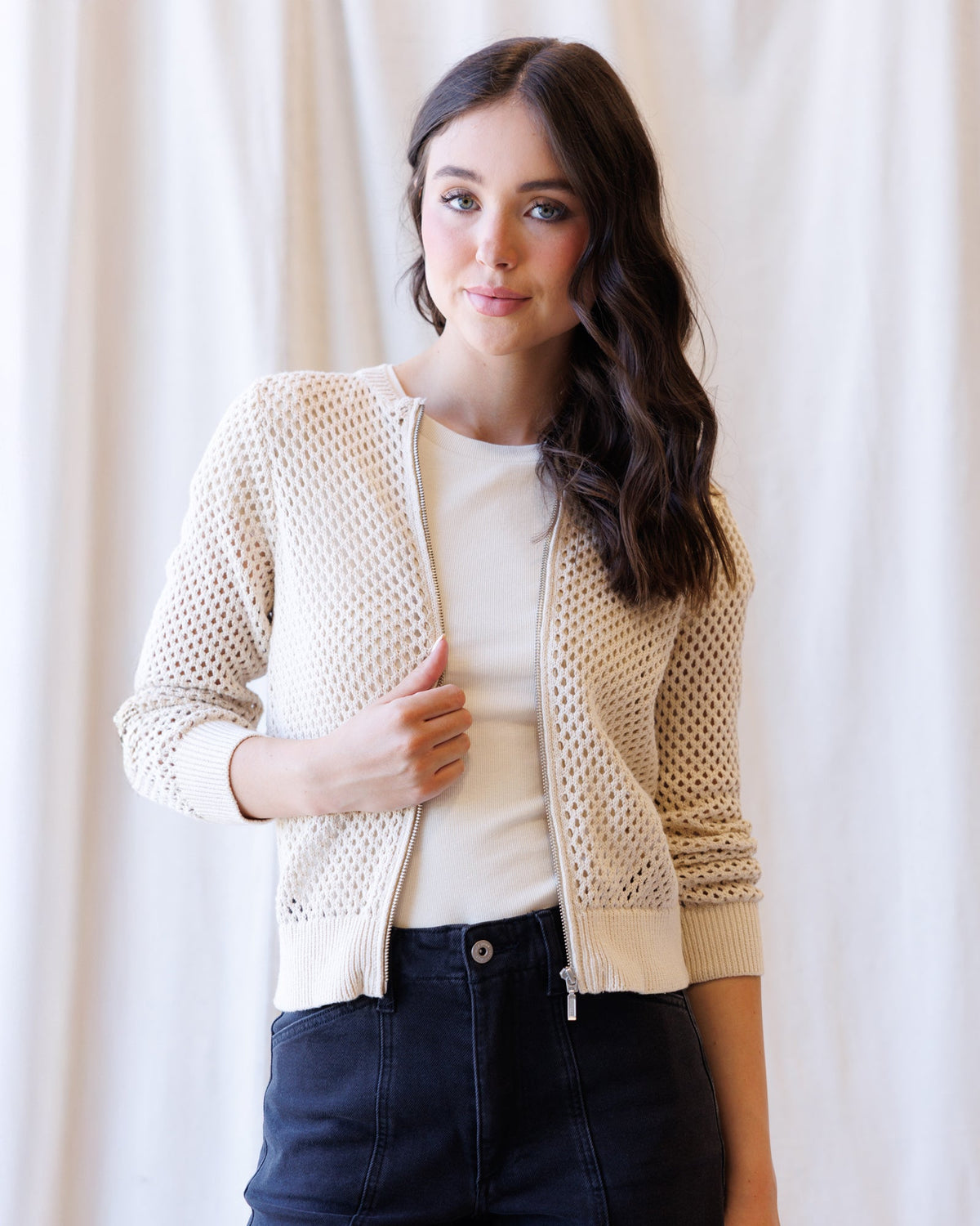 A woman wearing a cream-colored open-knit zip-up cardigan over a white top and dark jeans, standing against a soft white curtain background.