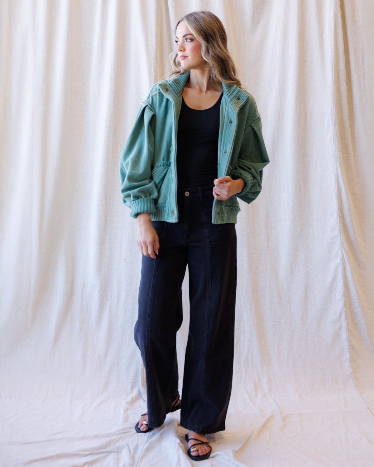 A woman modeling a sage green corduroy jacket over a black top and black wide-leg pants, standing against a draped cream fabric backdrop.