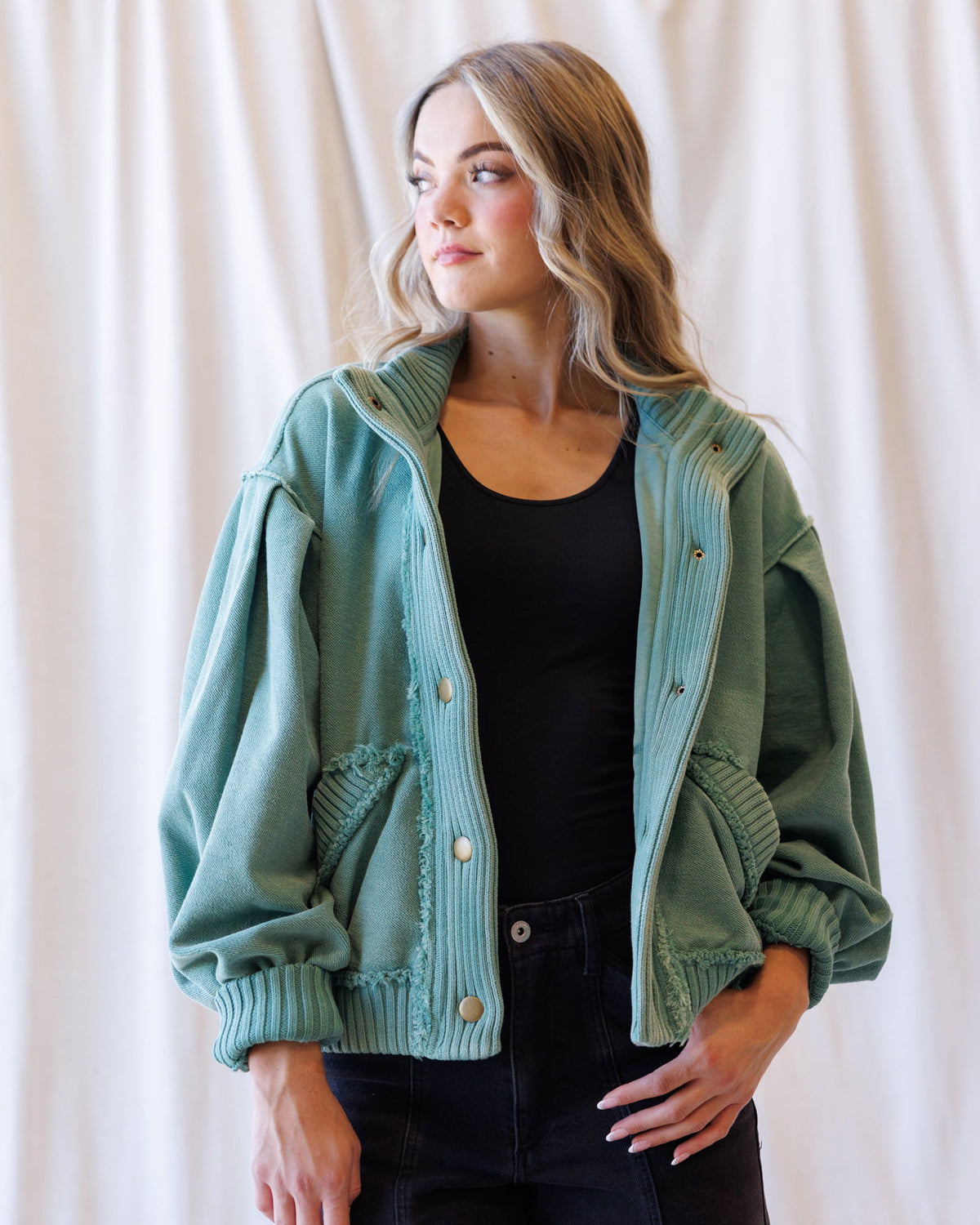 A woman wearing a sage green corduroy jacket with button details and ribbed trim, layered over a black top and dark jeans, standing against a draped white fabric background.