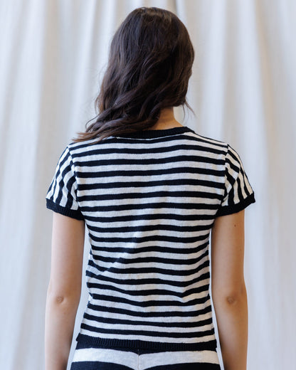A woman viewed from behind wearing a short-sleeve knit top with horizontal black and white stripes, set against a soft cream-colored curtain background.