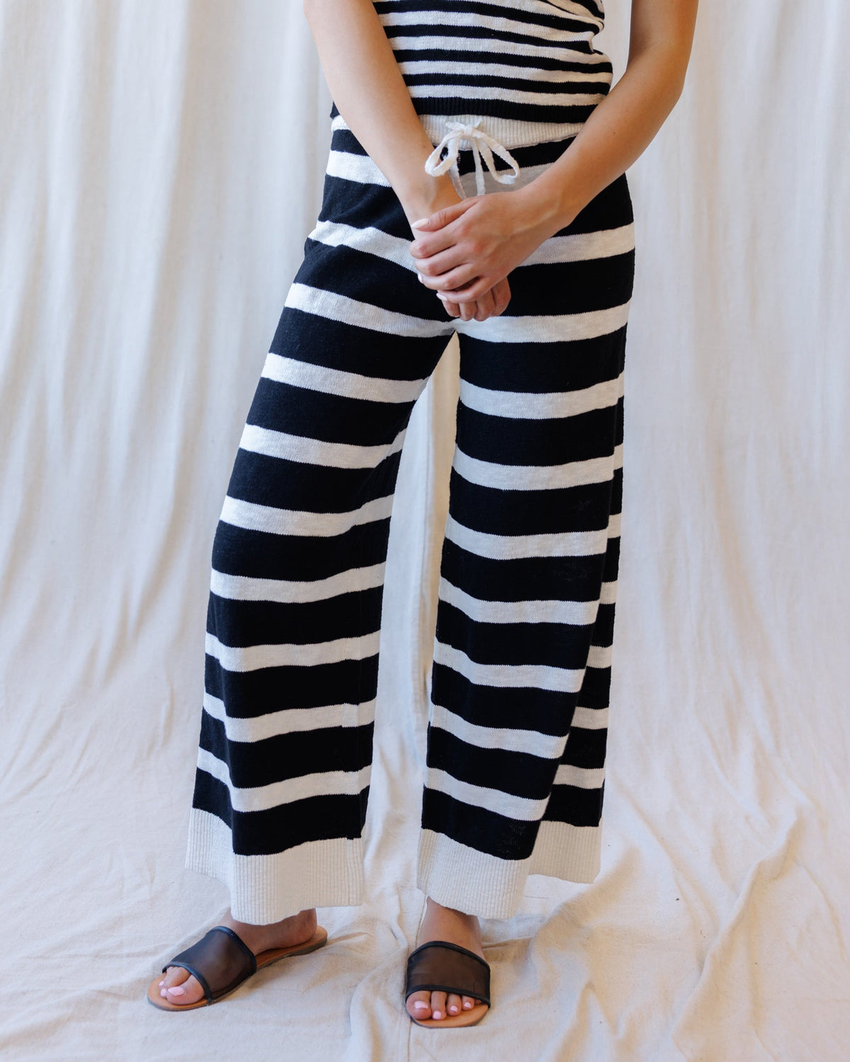 A person wearing black and white horizontally striped wide-leg knit pants with a drawstring waist, paired with dark brown slide sandals, standing against a draped white fabric backdrop.